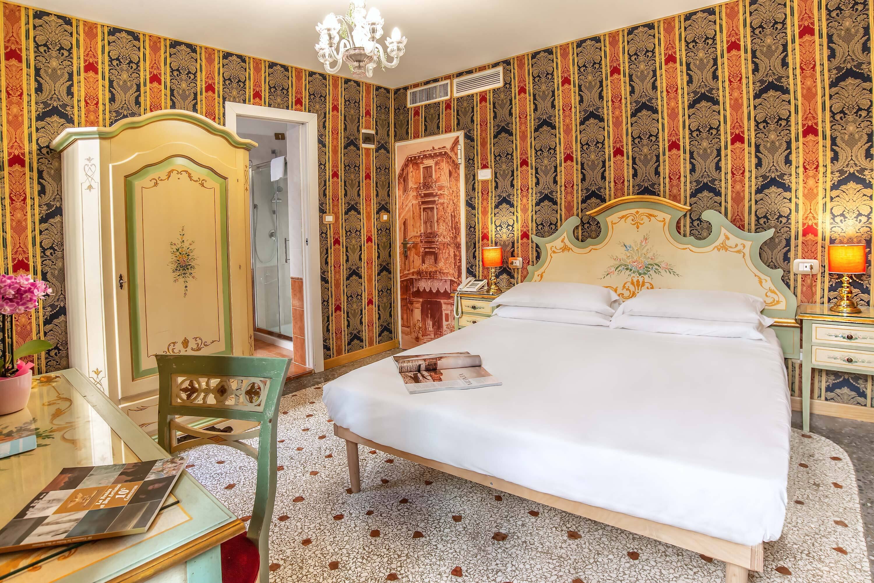 Ein Hotelzimmer mit einem großen Bett, einem Holzstuhl, einem Schreibtisch mit Lampe sowie einer Wand mit ornamentalem Wandpapier.
