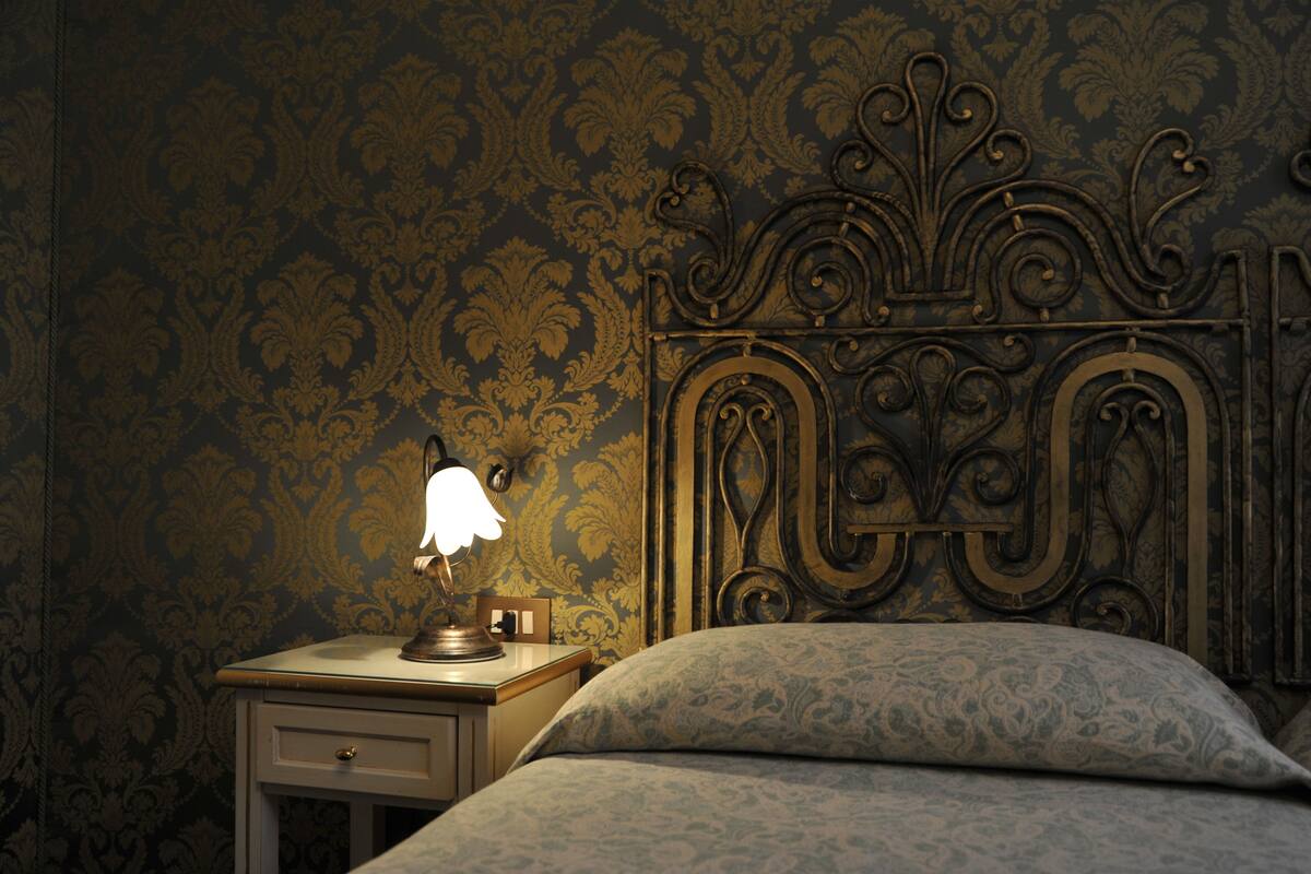 Une chambre à coucher avec une tête de lit de style vintage, une table de chevet avec une lampe et un mur orné d’un motif.