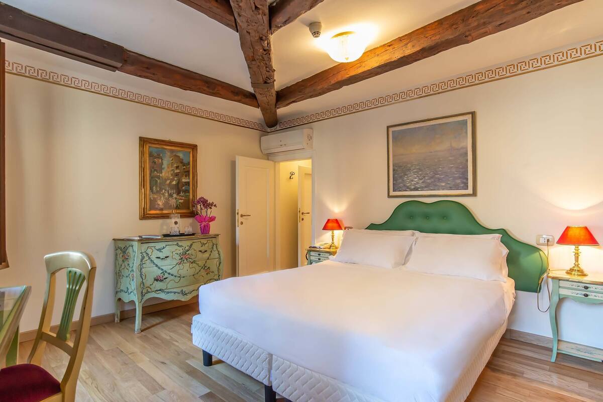 Un lit bien fait, avec du linge de lit blanc, une tête de lit recouverte d’un tissu vert et deux lampes de chevet.