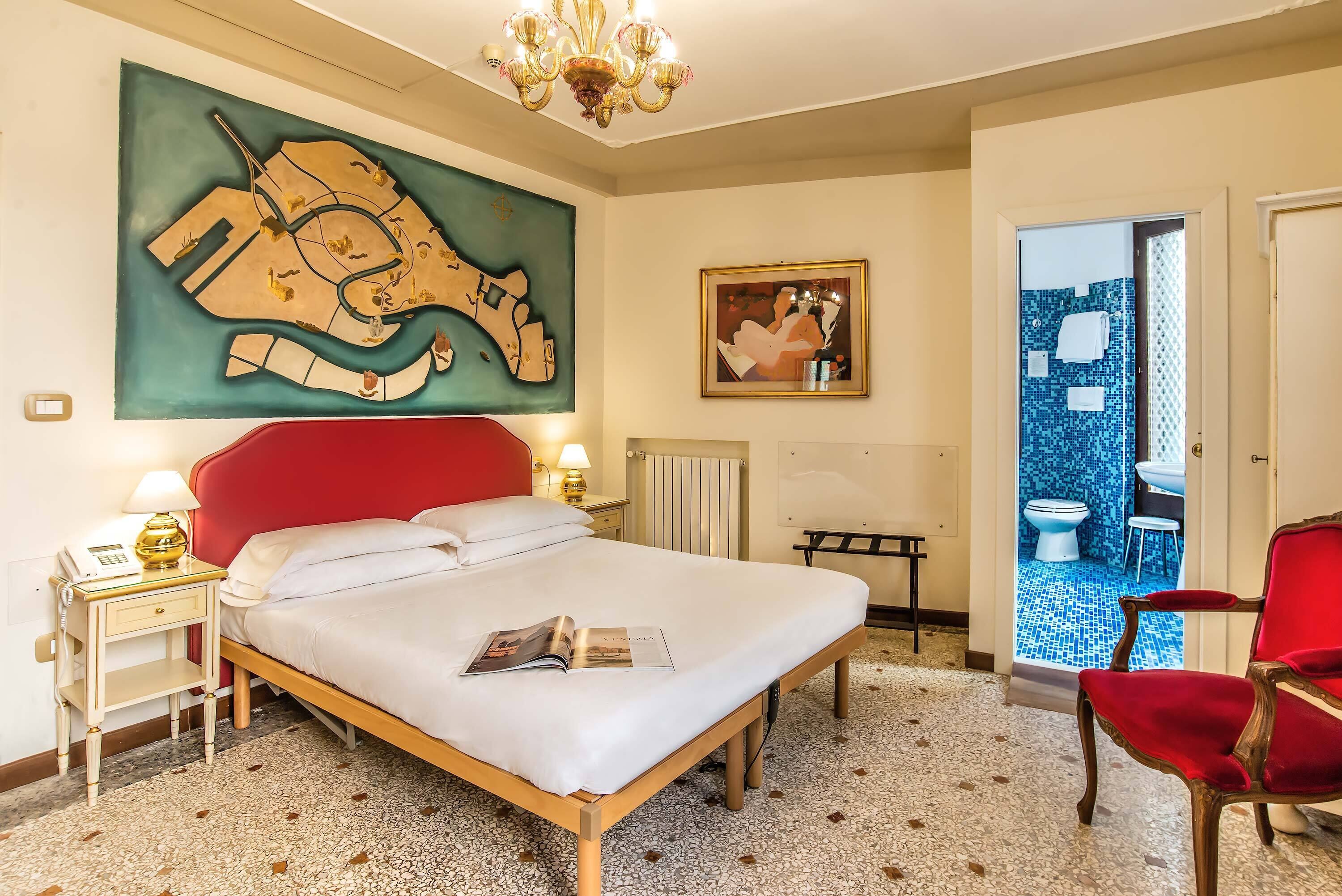 A hotel room with a bed, a red chair, a map on the wall, a painting, and a bathroom visible through an open door.