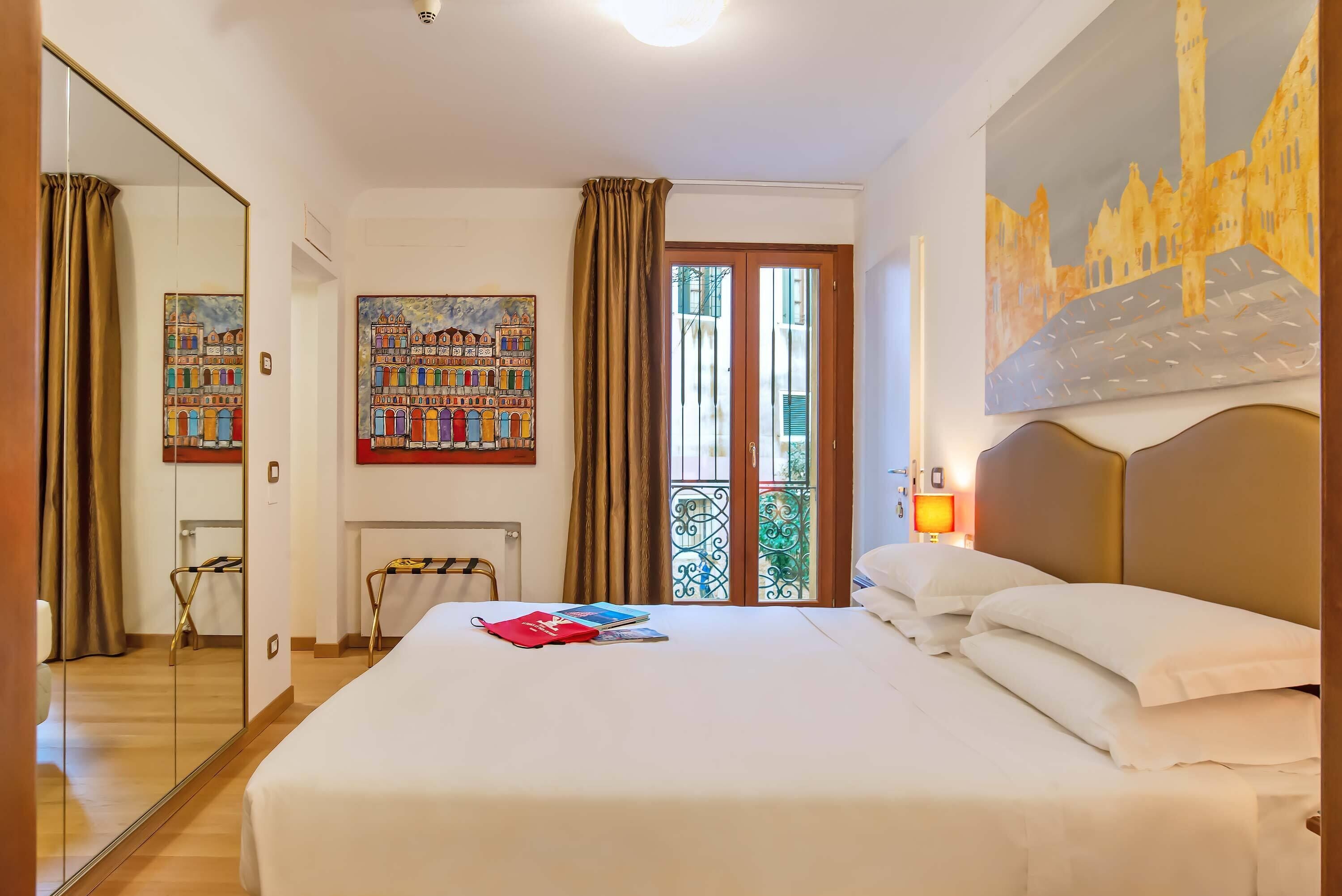 A hotel room with a large bed, a mirror, a painting, a view of a cityscape, and a balcony with curtains.