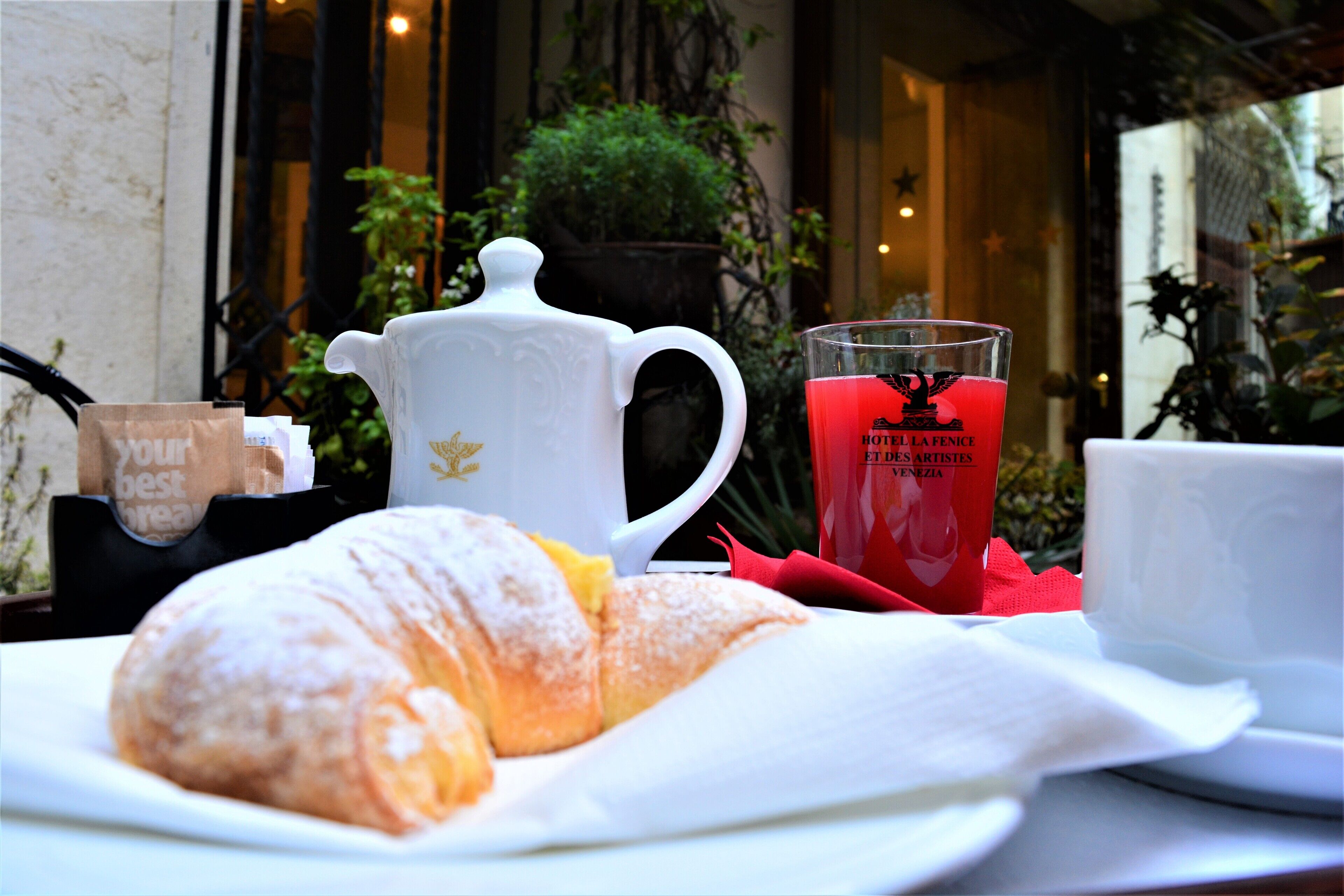 Un croissant en un plato, una tetera blanca, un vaso rojo y una bolsa de papel marrón con texto.