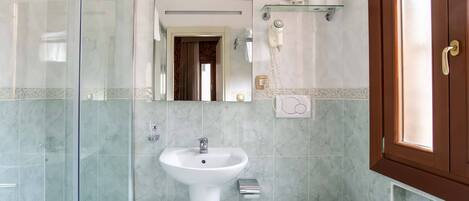 A bathroom with a glass shower enclosure, a pedestal sink, a toilet, and a hairdryer on a shelf.