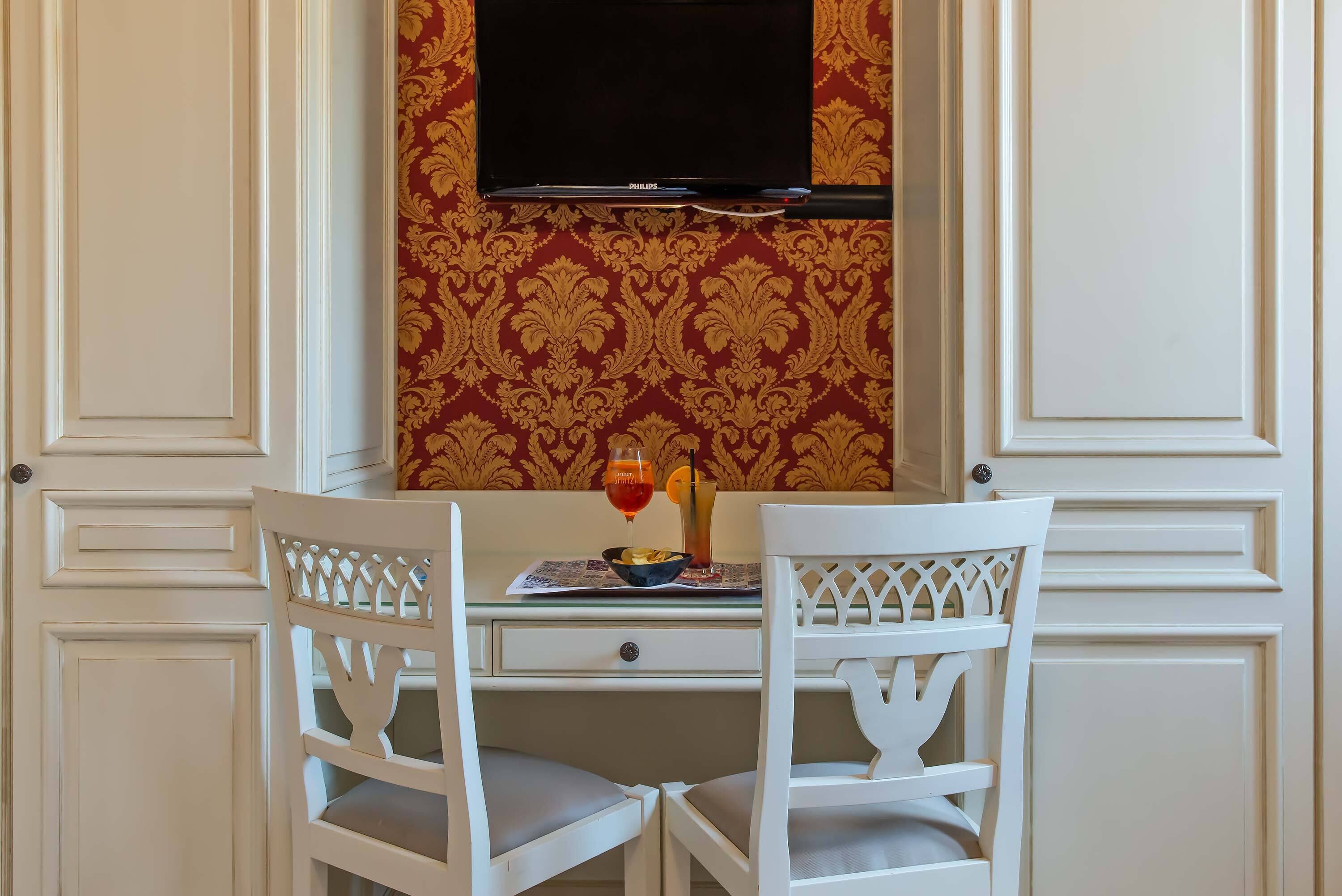 Una habitación con una mesa blanca, dos sillas blancas, un televisor de pantalla plana montado encima de la mesa, y un papel tapiz estampado.