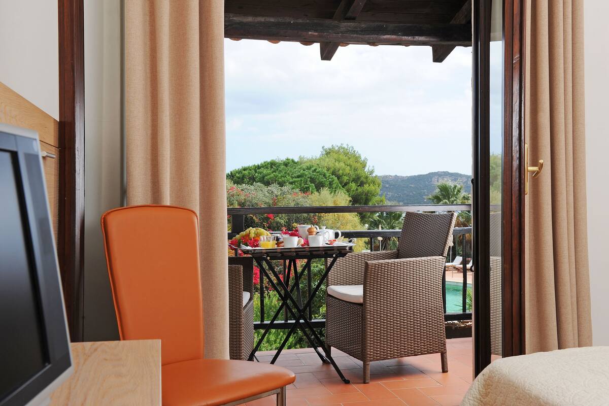 A balcony with wicker furniture, a table with a fruit bowl, and a view of a pool and greenery.