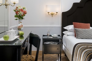 A hotel room with a bed, bedside table, a small desk with a chair, a vase with flowers, and a chandelier.