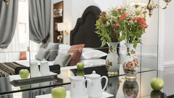 A hotel room with a bed, a bedside table with a vase of flowers, a green apple, and two white pitchers.