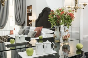 A hotel room with a bed, a bedside table with a vase of flowers, a green apple, and two white pitchers.