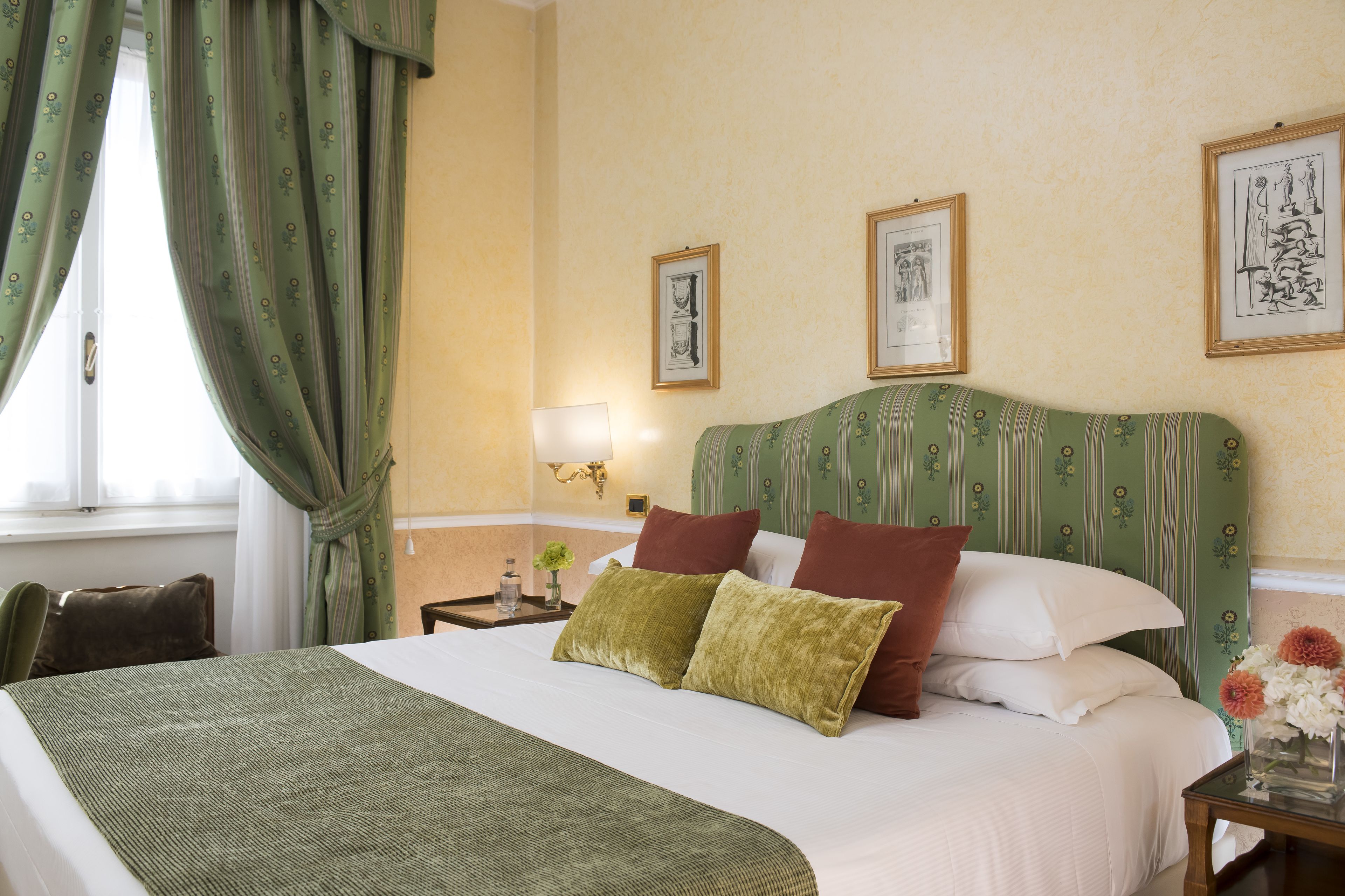 A neatly made bed with a green bedspread and pillows, a bedside table with a lamp, and framed artwork on the wall.