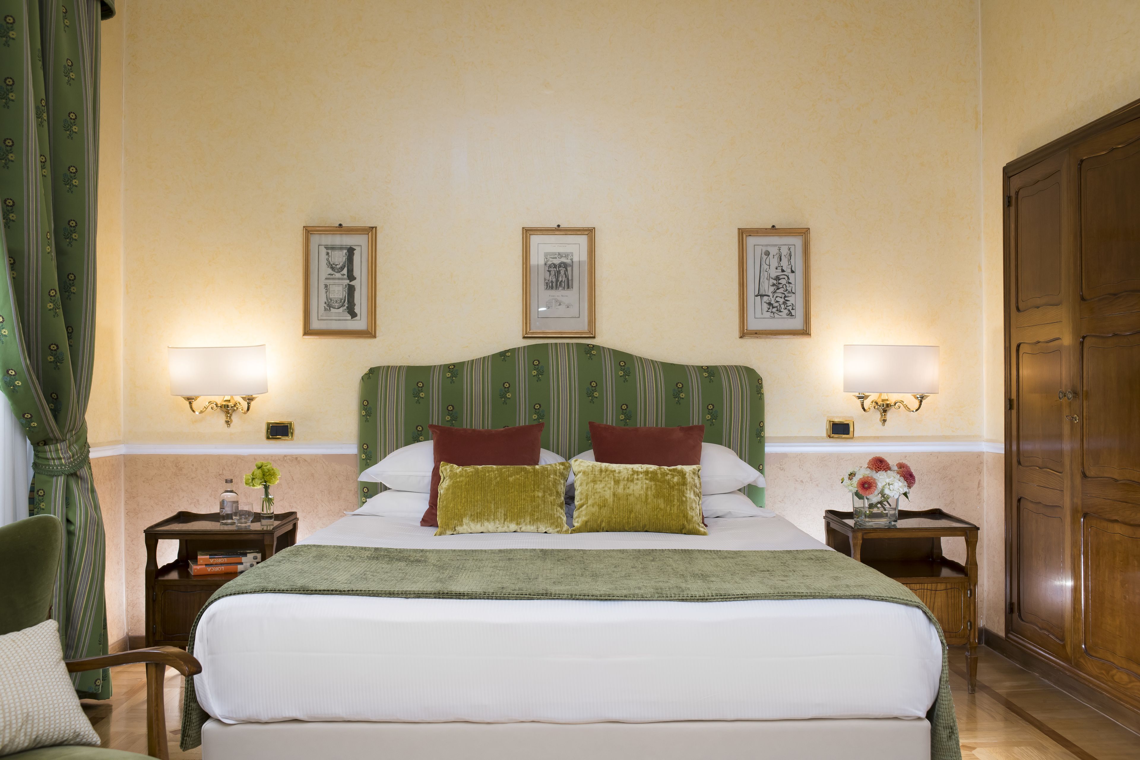 Una camera d'albergo con un letto grande, due lampade ai lati del letto, quadri appesi al muro, una testiera a righe verdi e un armadio in legno.