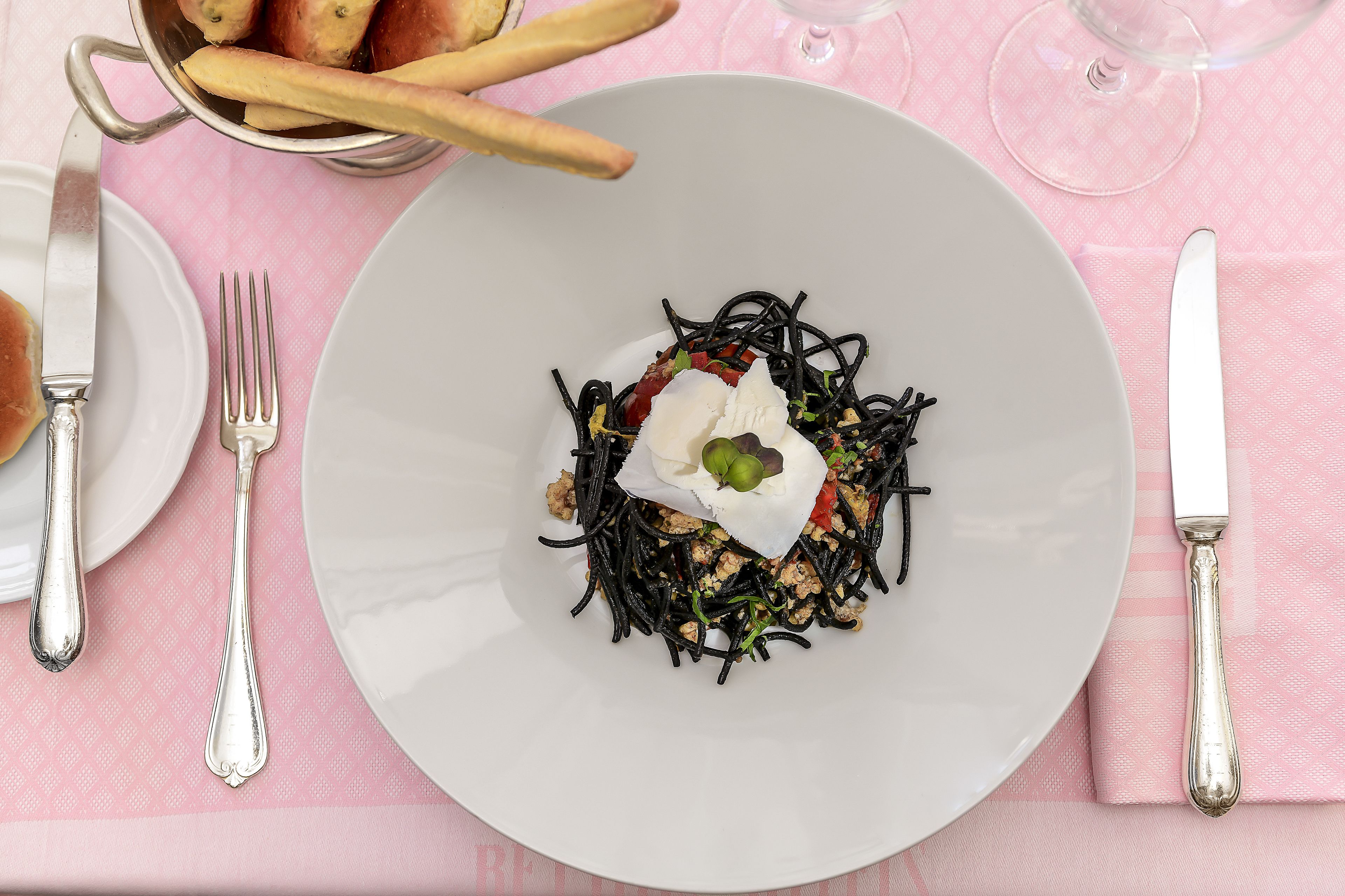 A plate of pasta with a dollop of sauce and a garnish, accompanied by a fork and knife on a pink tablecloth.