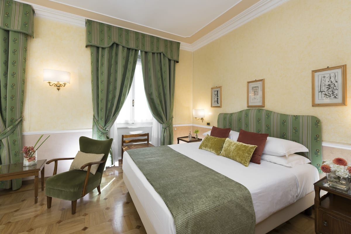 Ein Hotelzimmer mit einem großen Bett, einem Sessel, einem Tisch und einem Fenster mit Vorhängen.