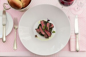 A plate of steak with greens and a glass of red wine on a pink tablecloth.