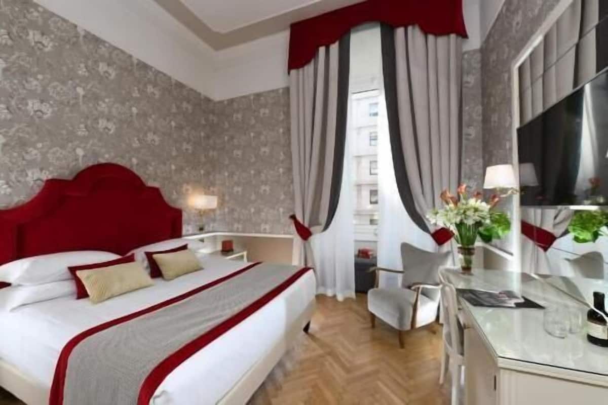 Ein Hotelzimmer mit einem großen Bett, einem Schreibtisch, einem Stuhl, einem Fernseher und einem Fenster mit Vorhängen.