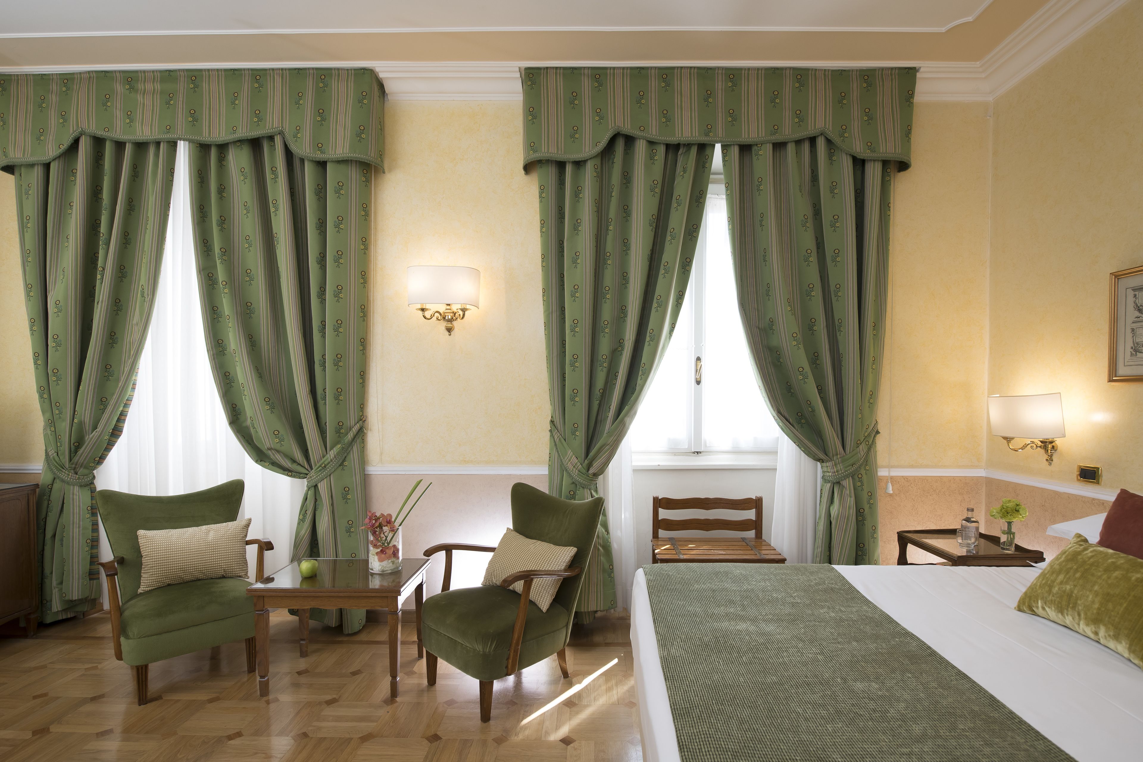 A hotel room with two green upholstered chairs, a wooden table, a bed with a green bedspread, and a nightstand with a lamp.