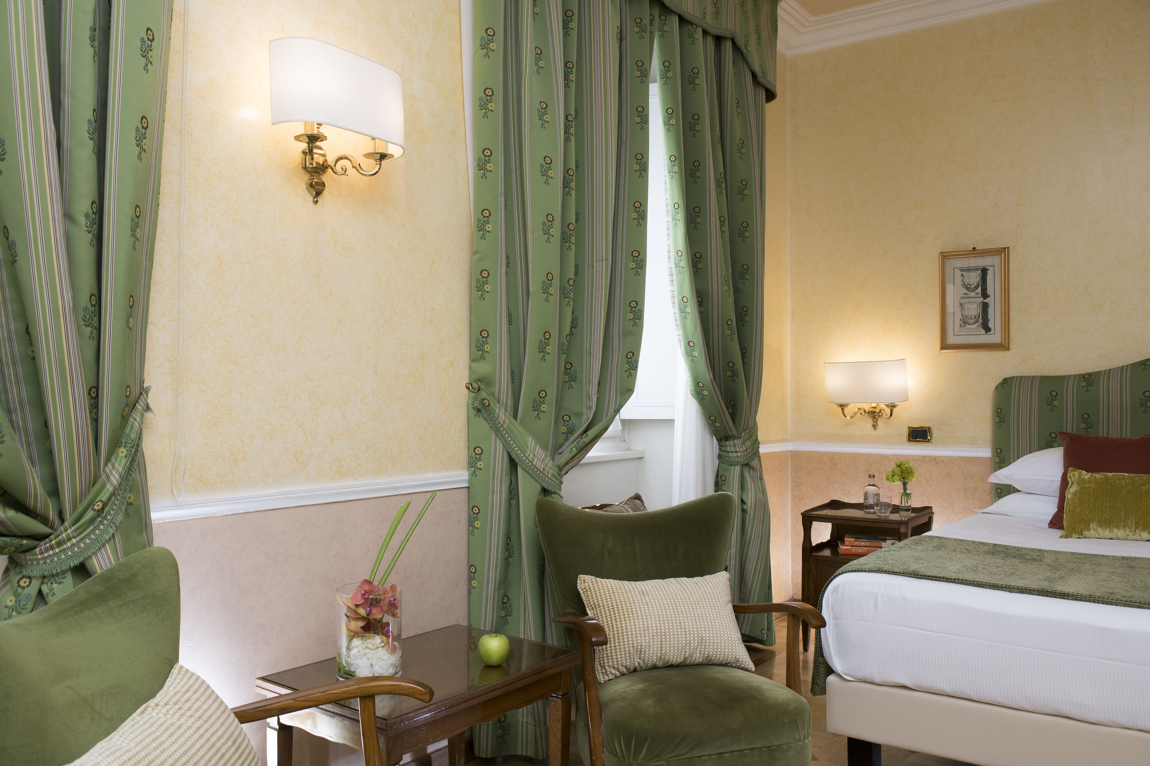 Une chambre d’hôtel avec un lit, une chaise, une table avec un vase et une pomme, et des rideaux à motifs floraux.