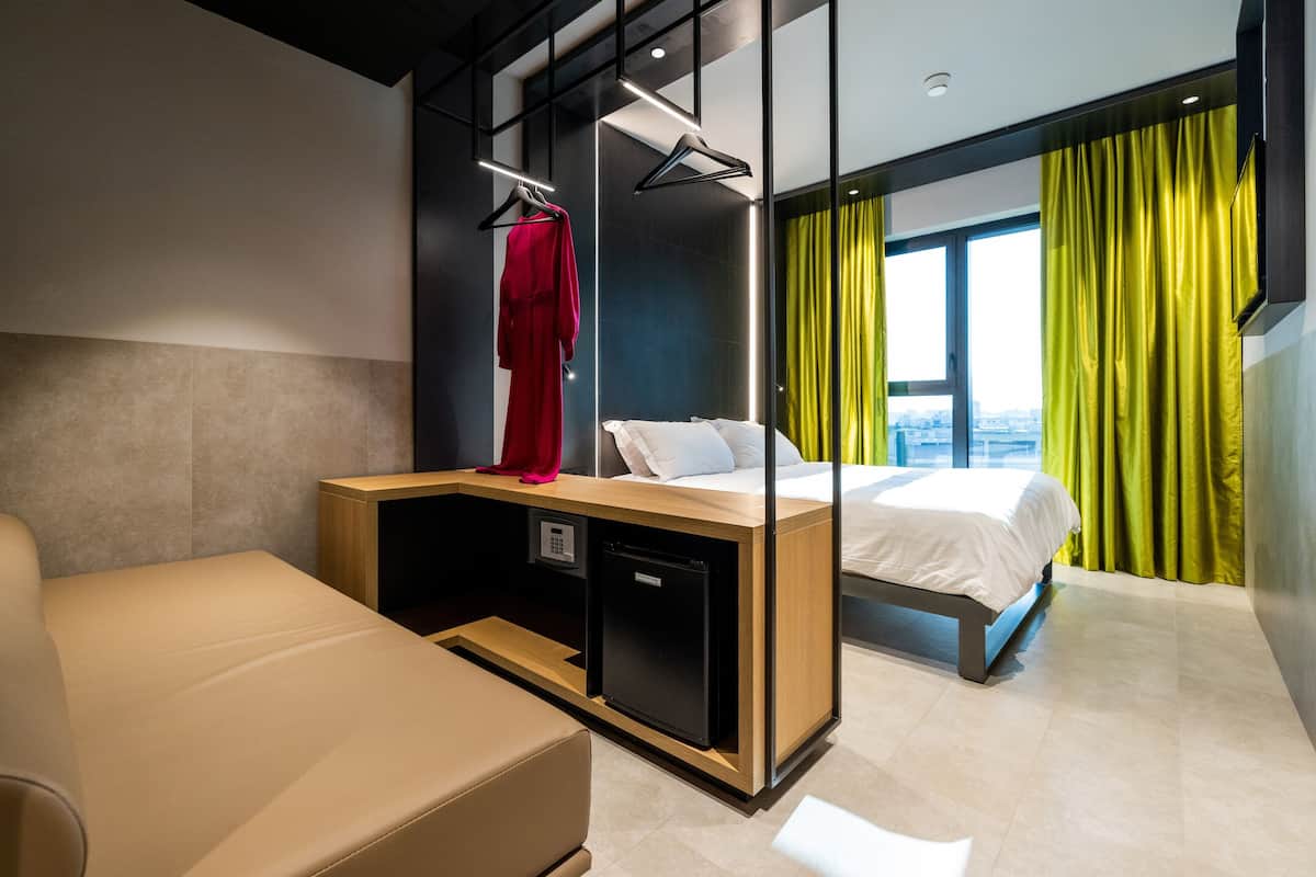Une chambre d’hôtel moderne équipée d’un lit, d’un bureau avec un ordinateur, d’un coin salon et offrant une vue sur la ville grâce à de grandes fenêtres.