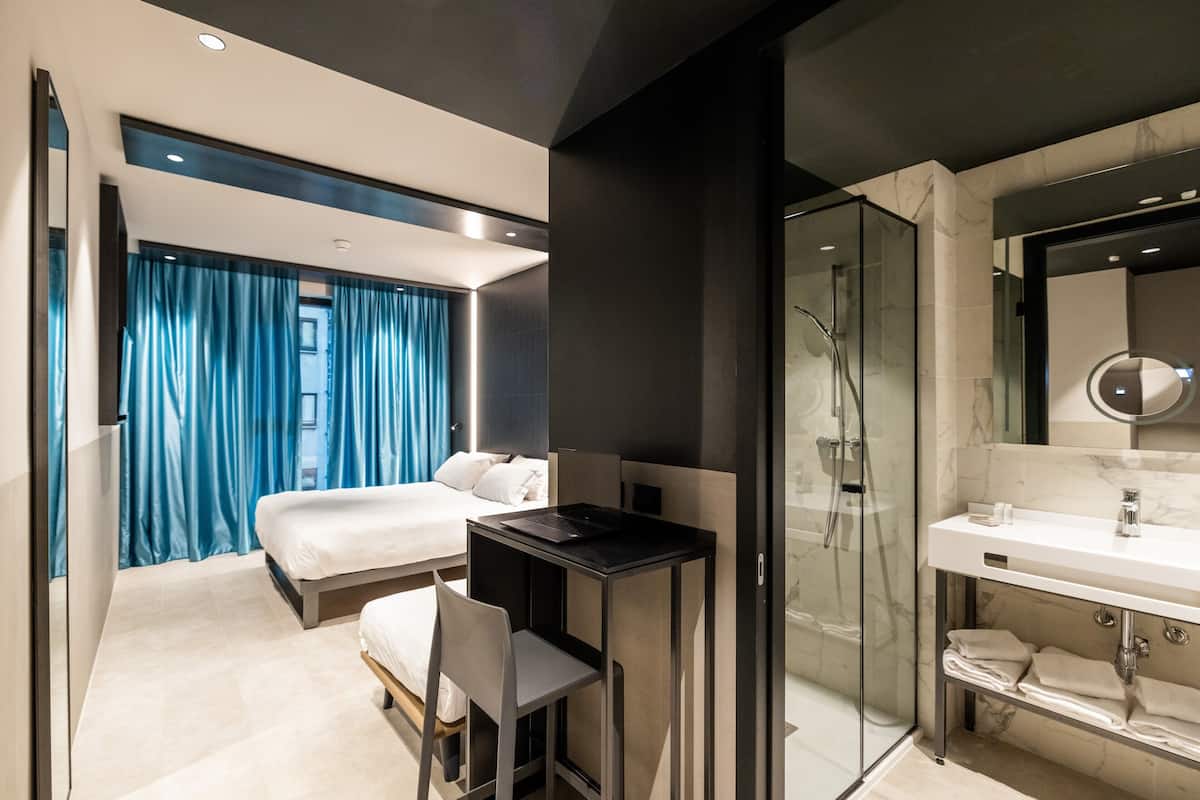 Une chambre d’hôtel moderne équipée d’un lit, d’un bureau, d’une douche et d’une salle de bain avec un miroir et un lavabo.