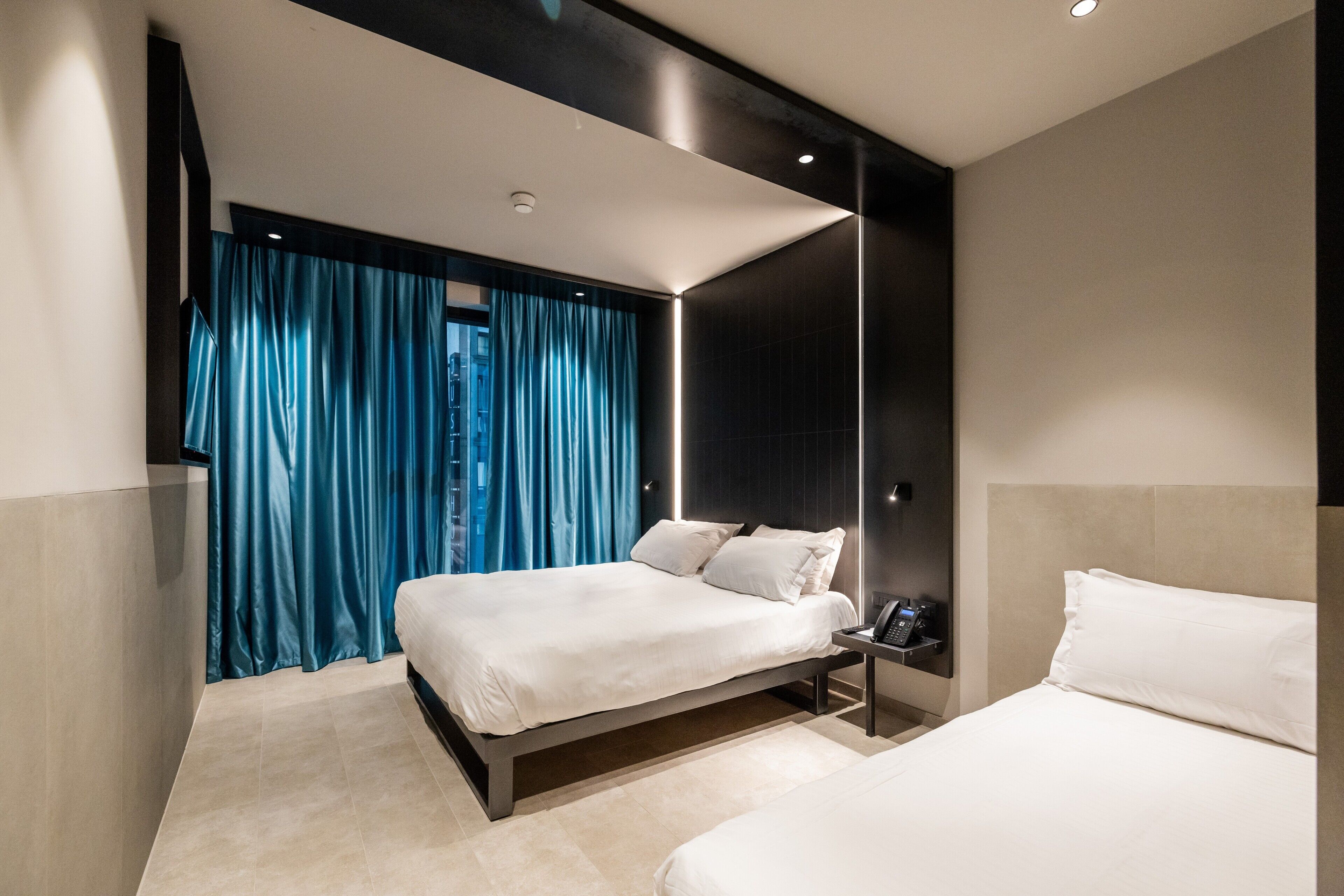 A modern hotel room with a large bed, a bedside table with a phone, and a view of the city through a window with blue curtains.