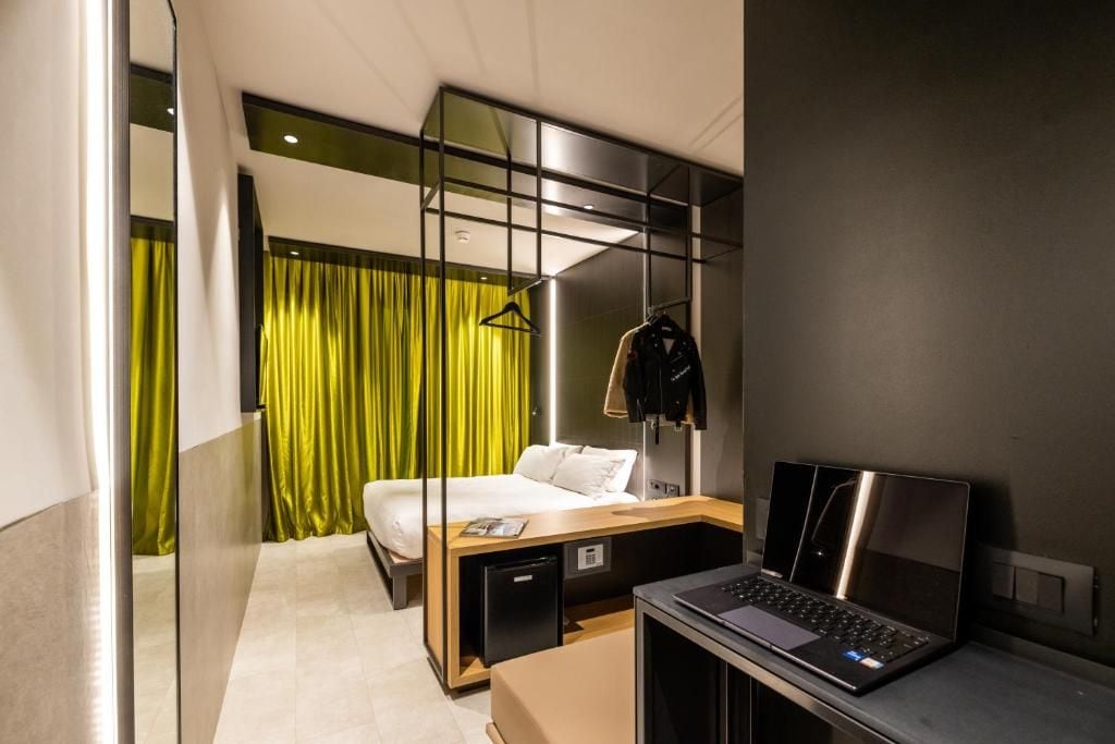 A modern hotel room with a bed, a desk with a laptop, a small cabinet, and a mirror.