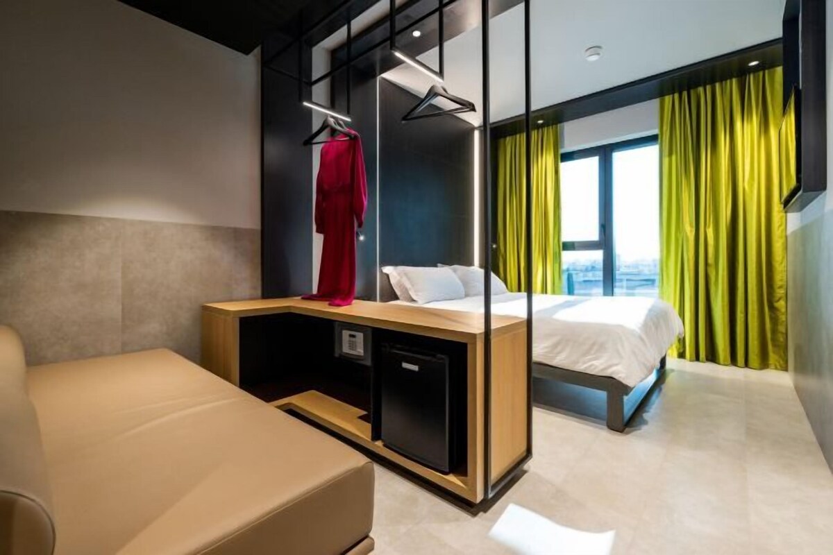A modern hotel room with a bed, a desk with a computer, a seating area, and a view of the city through large windows.