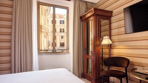 A hotel room with a large bed, a wooden cabinet, a chair, a small table with a lamp, and a window offering a view of buildings. - Mascagni Hotel (Rome)