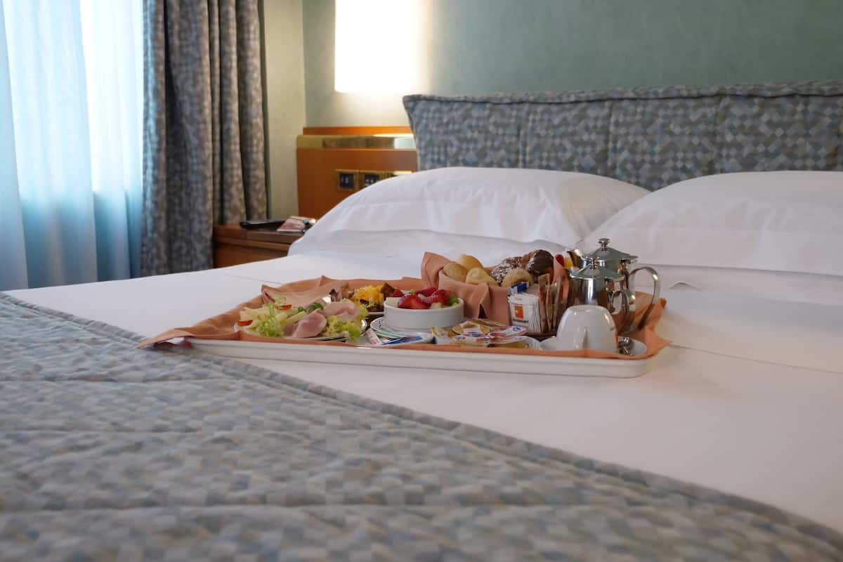 Habitación de hotel con una cama bien hecha, una bandeja con alimentos para el desayuno, una mesita de noche con una lámpara y una mesita auxiliar con un teléfono y un libro.