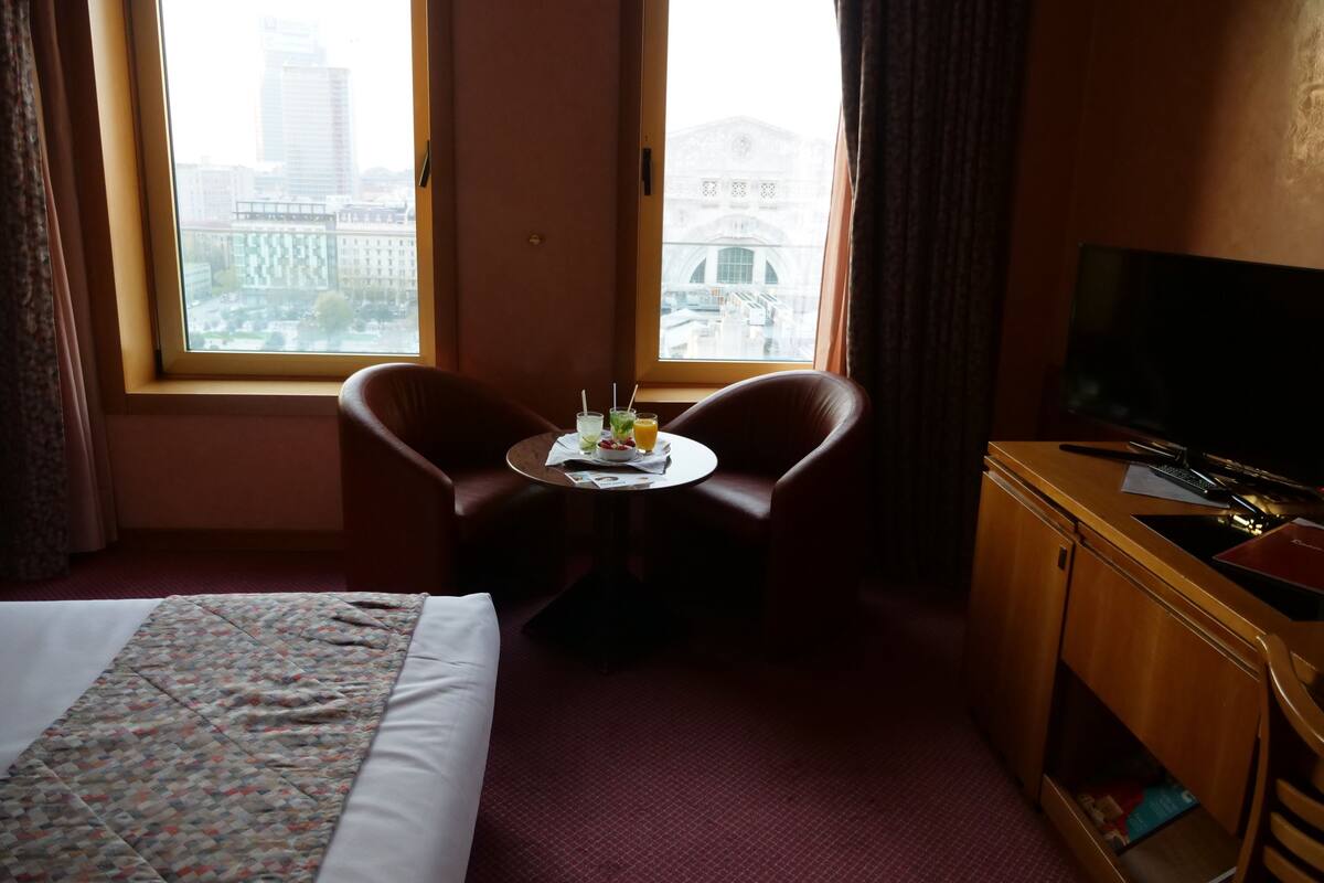 Habitación de hotel con una cama, un escritorio con televisor, una silla y una mesa con bebidas.
