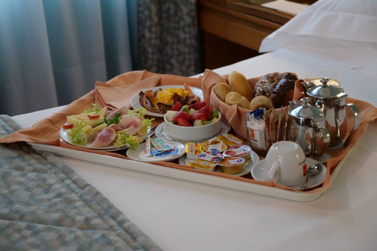 Bandeja de desayuno con diversas comidas, como frutas, pasteles y productos lácteos, sobre una cama en una habitación de hotel.