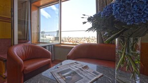 Une chambre d’hôtel avec vue sur le paysage urbain, visible à travers de grandes fenêtres, une table avec un journal et un vase rempli de fleurs.