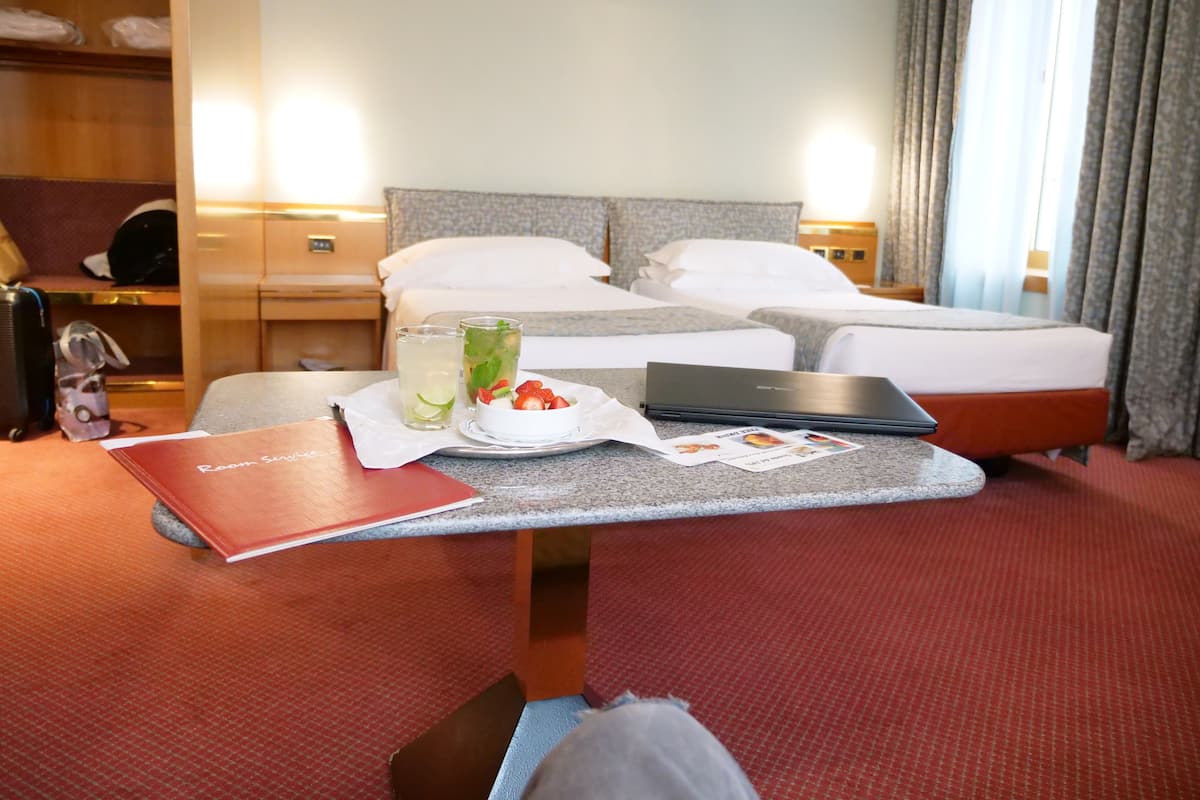 Habitación de hotel con una cama, un escritorio con una computadora portátil, una bandeja con una bebida y fruta, y una carpeta roja con la etiqueta 'Servicio a la habitación'.