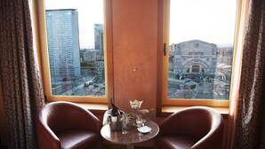 Une pièce avec deux fauteuils en cuir, une petite table sur laquelle se trouvent une bouteille, des verres et une assiette de nourriture. Une grande fenêtre offre une vue sur un paysage urbain avec des immeubles et un bâtiment historique.