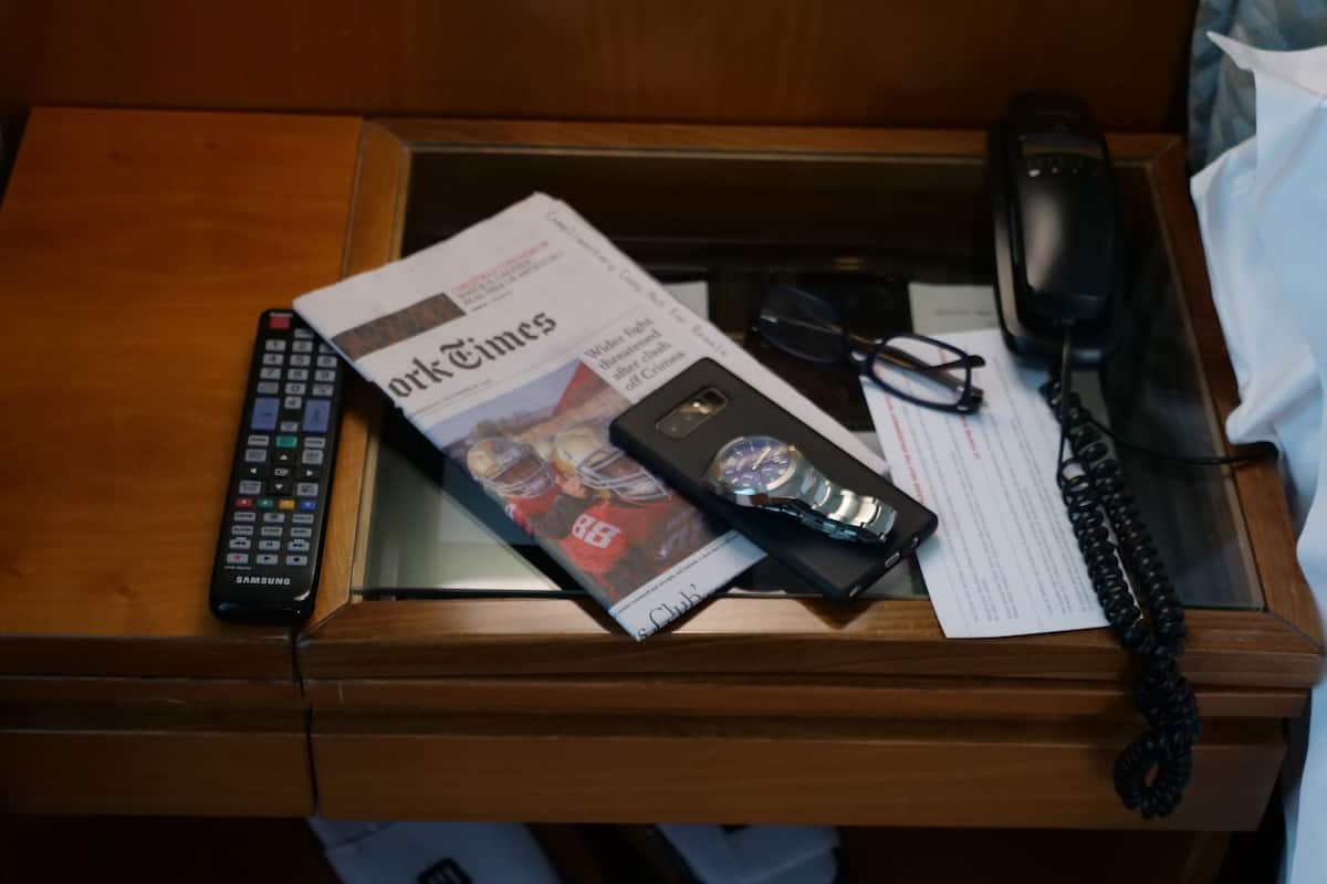 Un escritorio de madera con un control remoto, un periódico, un reloj, gafas, un teléfono y un teléfono alámbrico negro.