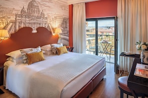 A hotel room with a large bed, a desk with a chair, a balcony with a table and chairs, and a mural of a famous building on the wall. - Starhotels Michelangelo (Rome)