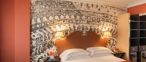 Habitación de hotel con un gran tapiz en la pared, una cama con sábanas blancas, dos lámparas de noche y una mesita con libros y un jarrón de flores.