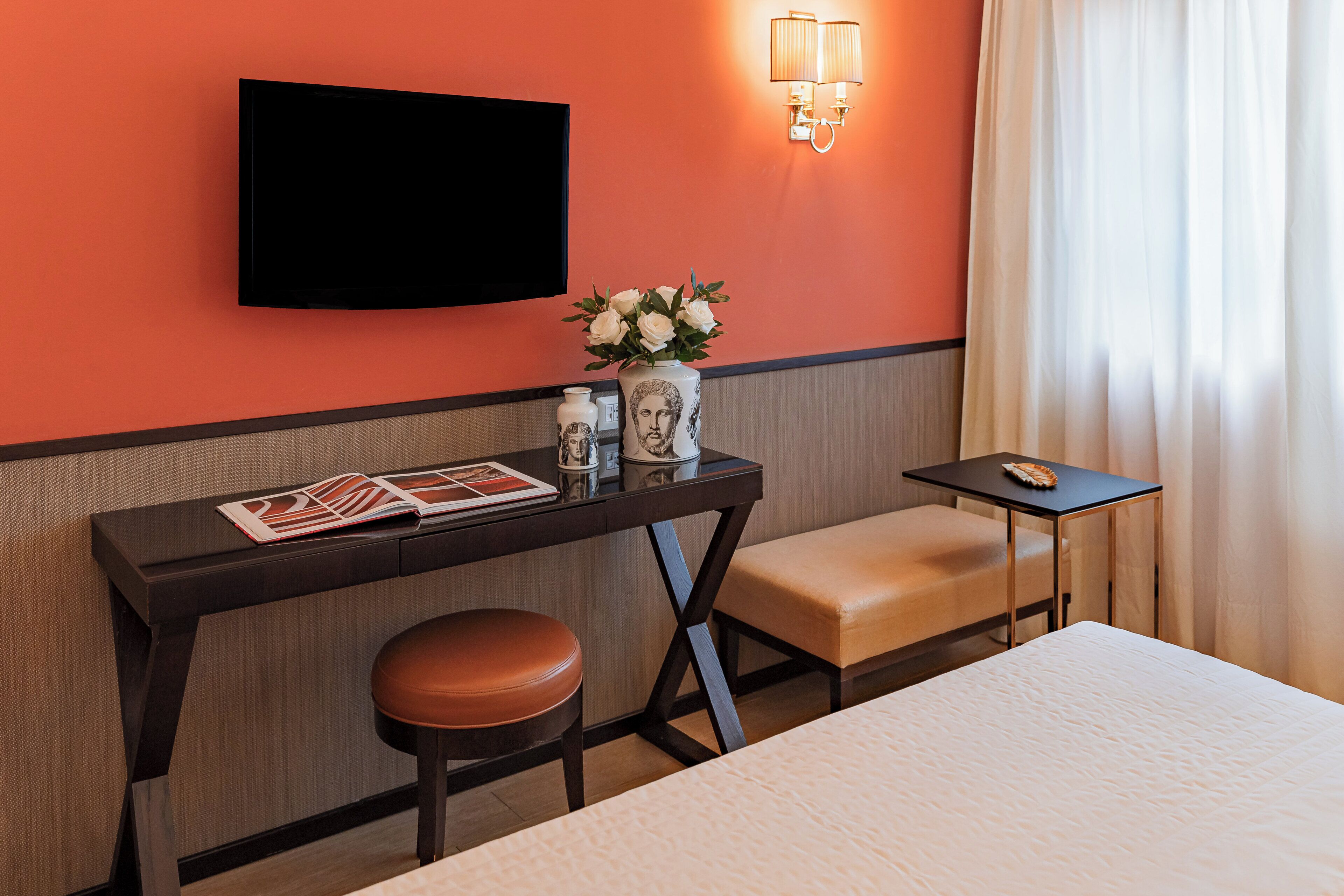 A hotel room with a bed, a desk with a chair, a TV mounted on the wall, a vase with flowers, and a small table with a lamp.