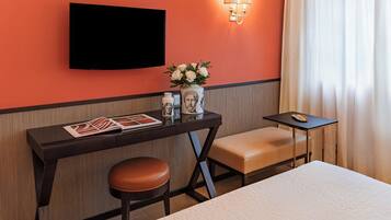 A hotel room with a bed, a desk with a chair, a TV mounted on the wall, a vase with flowers, and a small table with a lamp.