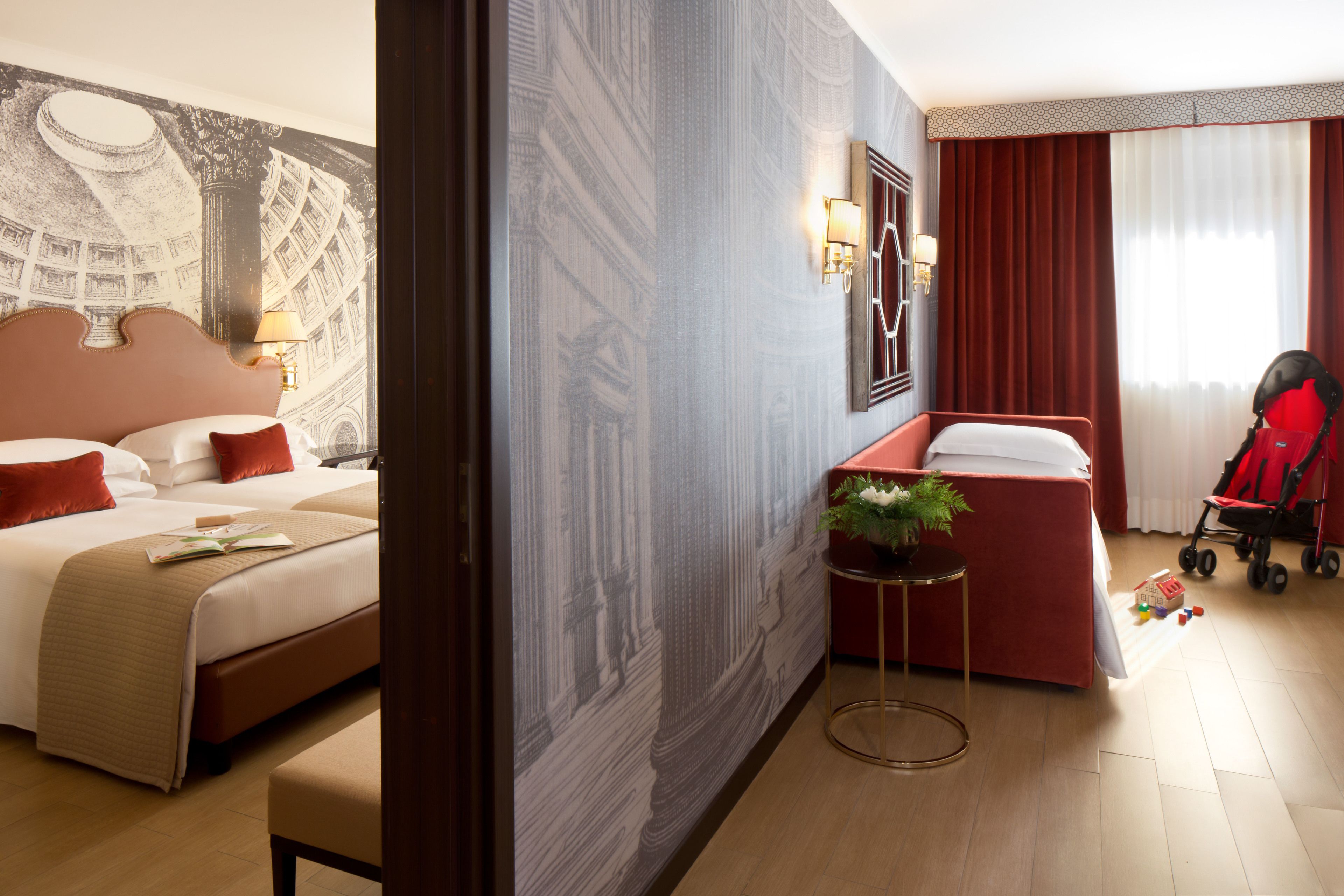 A hotel room with a bed, a desk, a red chair, a stroller, and a wall with a patterned mural.