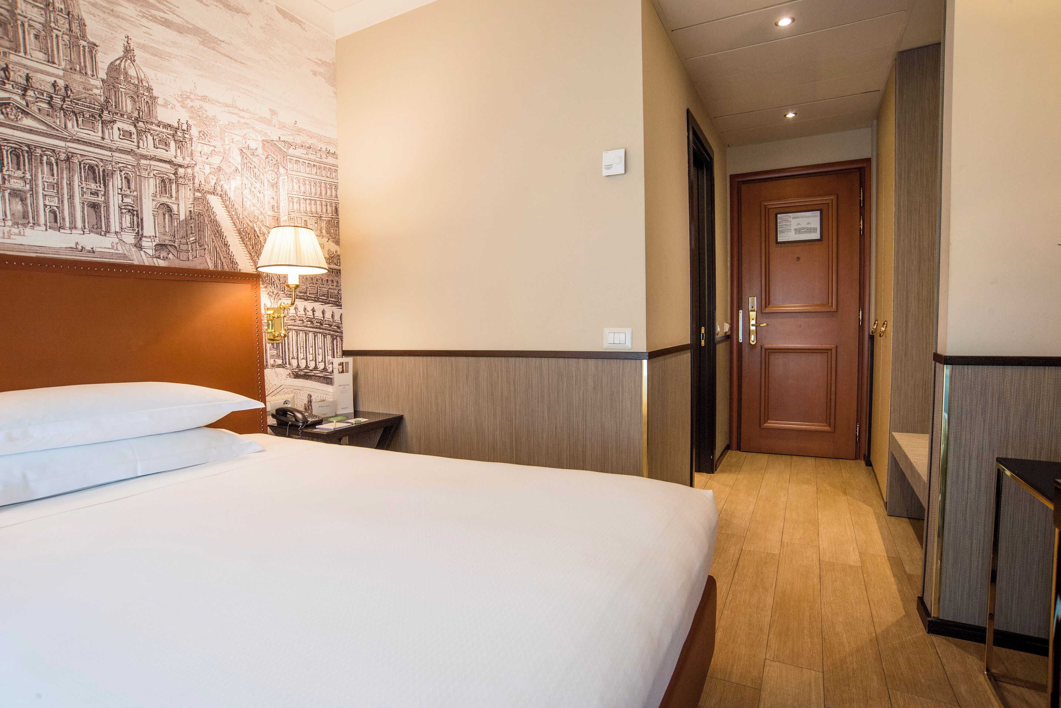 Une chambre d’hôtel avec un grand lit, une table de chevet, une lampe, une fresque murale et une porte donnant sur une autre pièce.