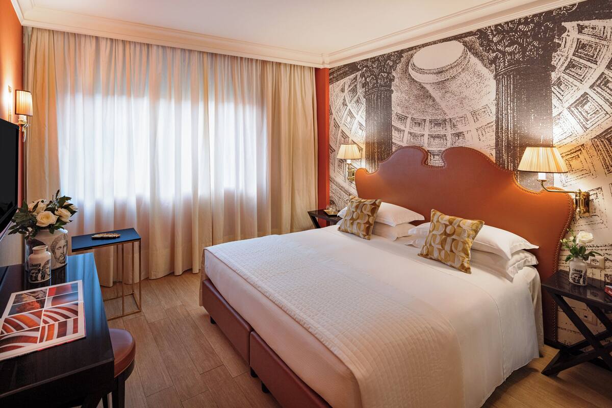 A hotel room with a large bed, a desk with a chair, a television, and a wall with a cityscape mural.