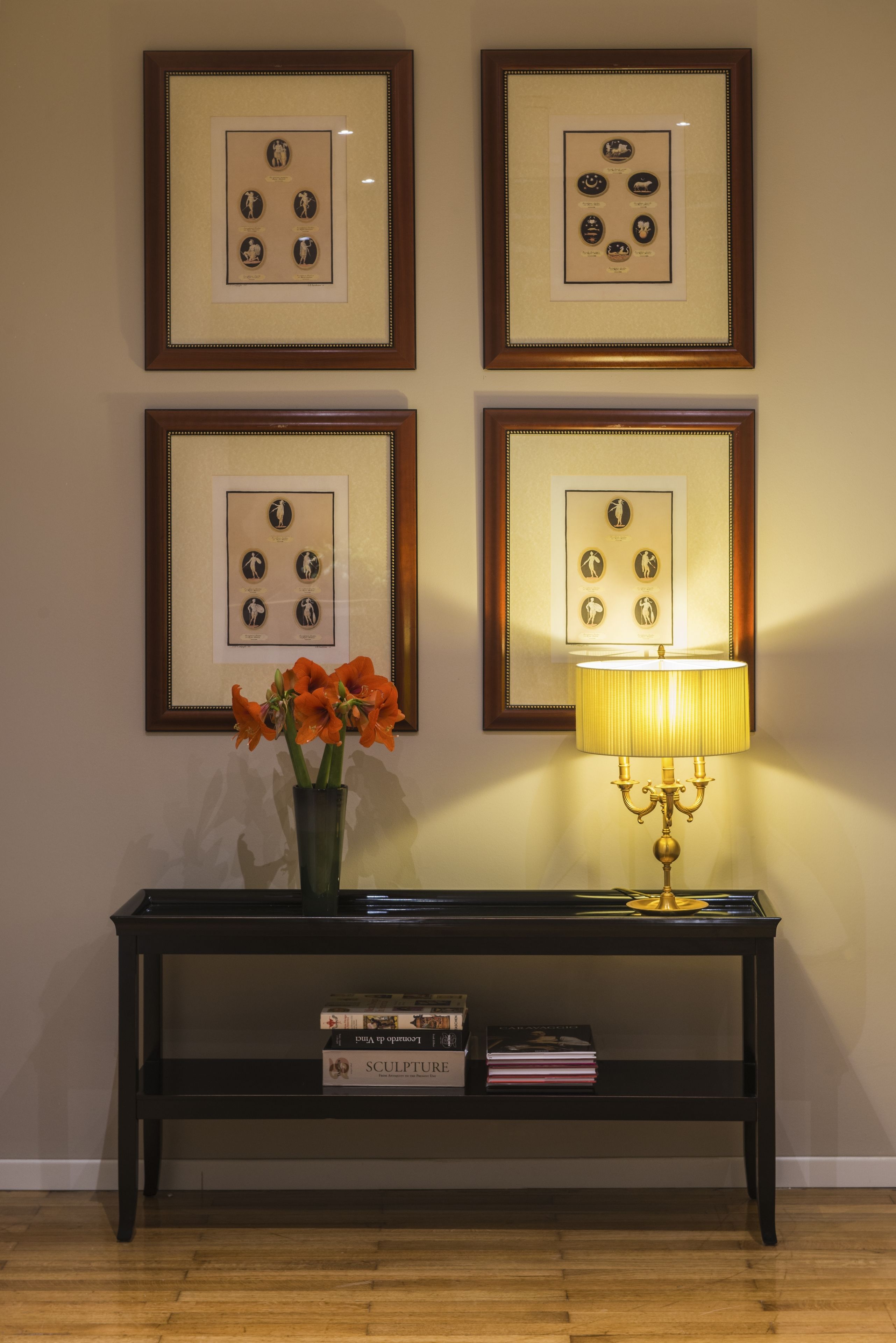A wall with four framed pictures, a table with a lamp, and a vase with flowers.
