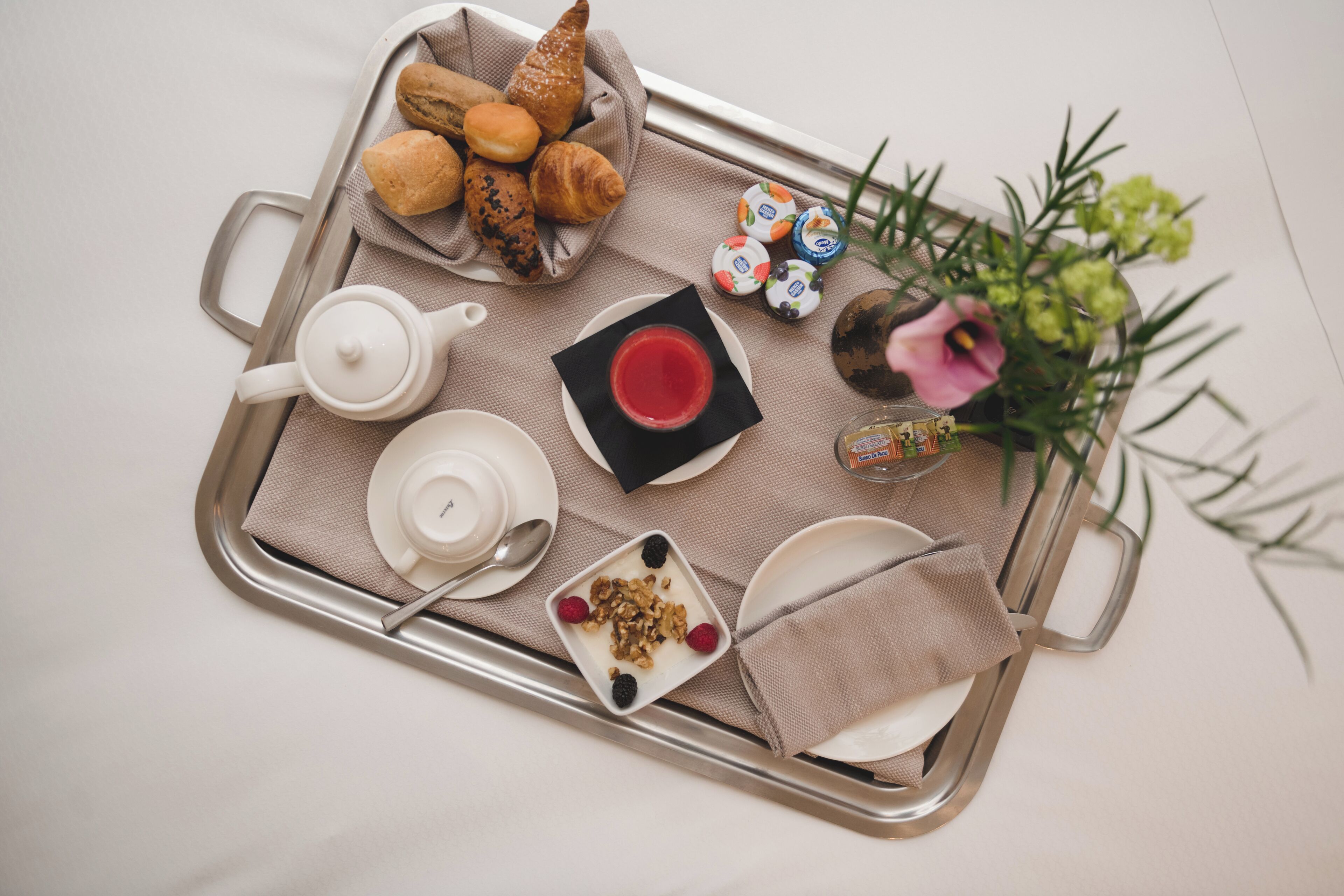 A tray with croissants, a teapot, a cup of tea, a bowl of granola with berries, and a small vase with flowers.