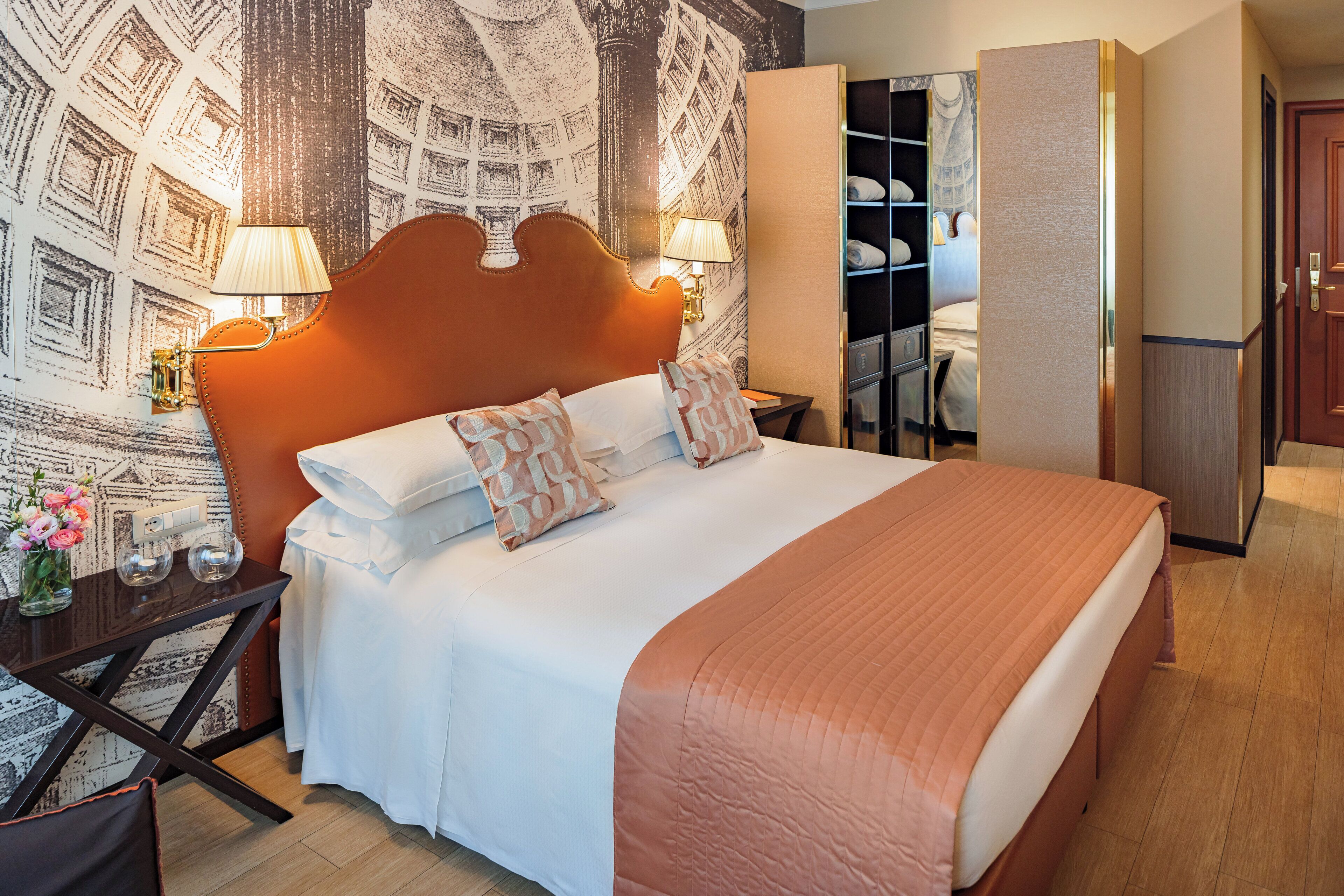 Een keurig opgeruimde hotelkamer met een groot bed, nachtlampjes, een klein tafeltje met een bloemenvaas en een kledingkast.