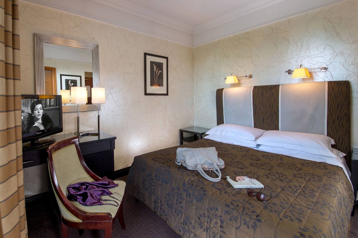 A hotel room with a bed, a chair, a desk with a television, and a lamp.