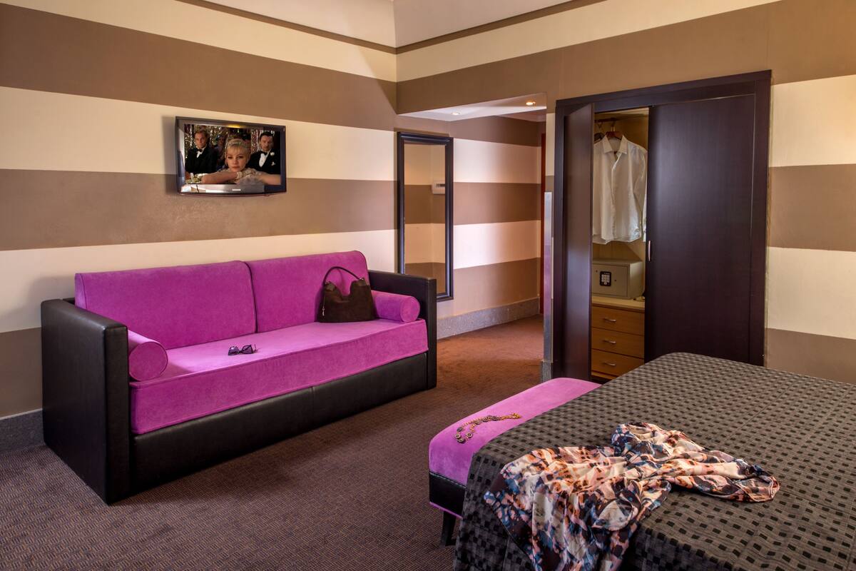 A hotel room with a purple sofa, a bed with a patterned blanket, a mirror, and a television on the wall.