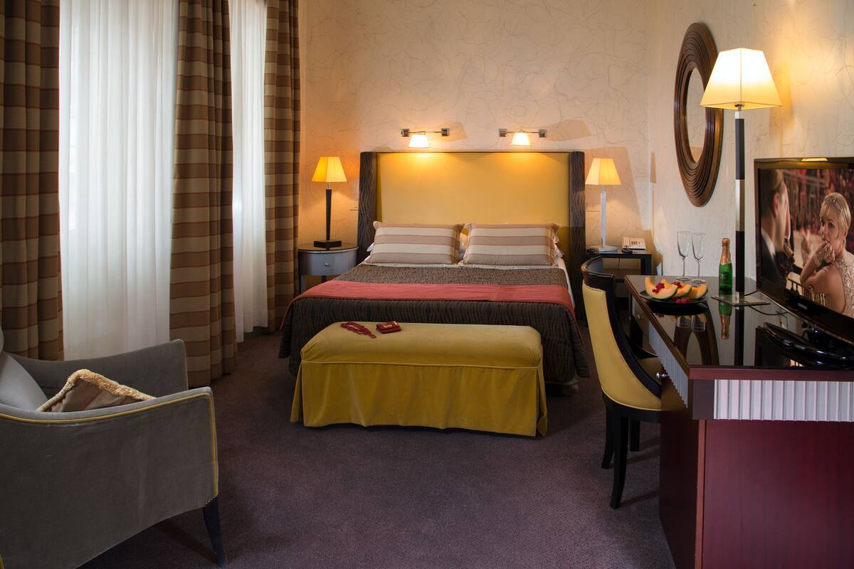 A hotel room with a bed, bedside lamps, a desk with a chair, a television, and a small table with fruit and glasses.