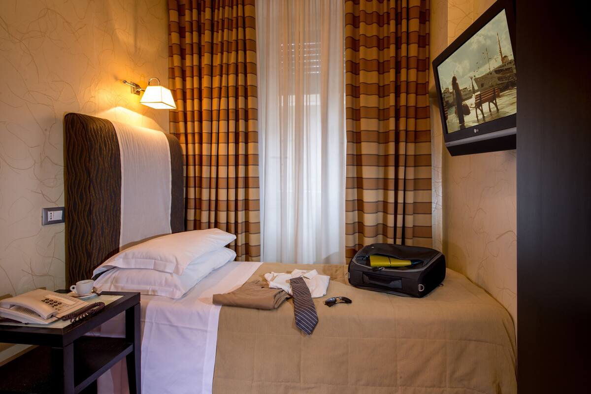 A hotel room with a bed, a desk with a telephone and a cup, a suitcase, and a television on the wall.
