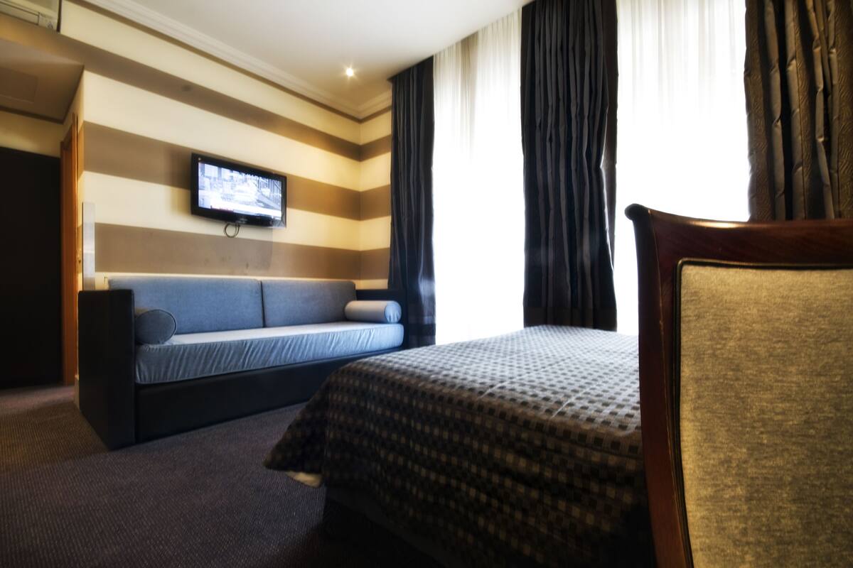 A hotel room with a bed, a sofa, a TV mounted on the wall, and a window with curtains.