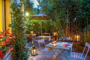 Een terras buiten met tafels en stoelen, omgeven door weelderige begroeiing en bloeiende planten.