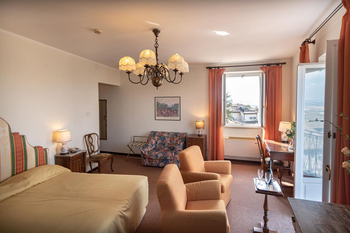 A hotel room with a bed, two armchairs, a small table with glasses, a lamp, a chair, a painting, and a view through the window.